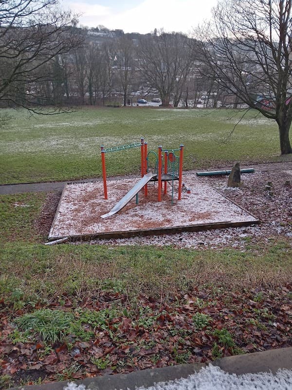 Playground with a slide and climbing frame, surrounded by grass and trees, lightly dusted with snow.