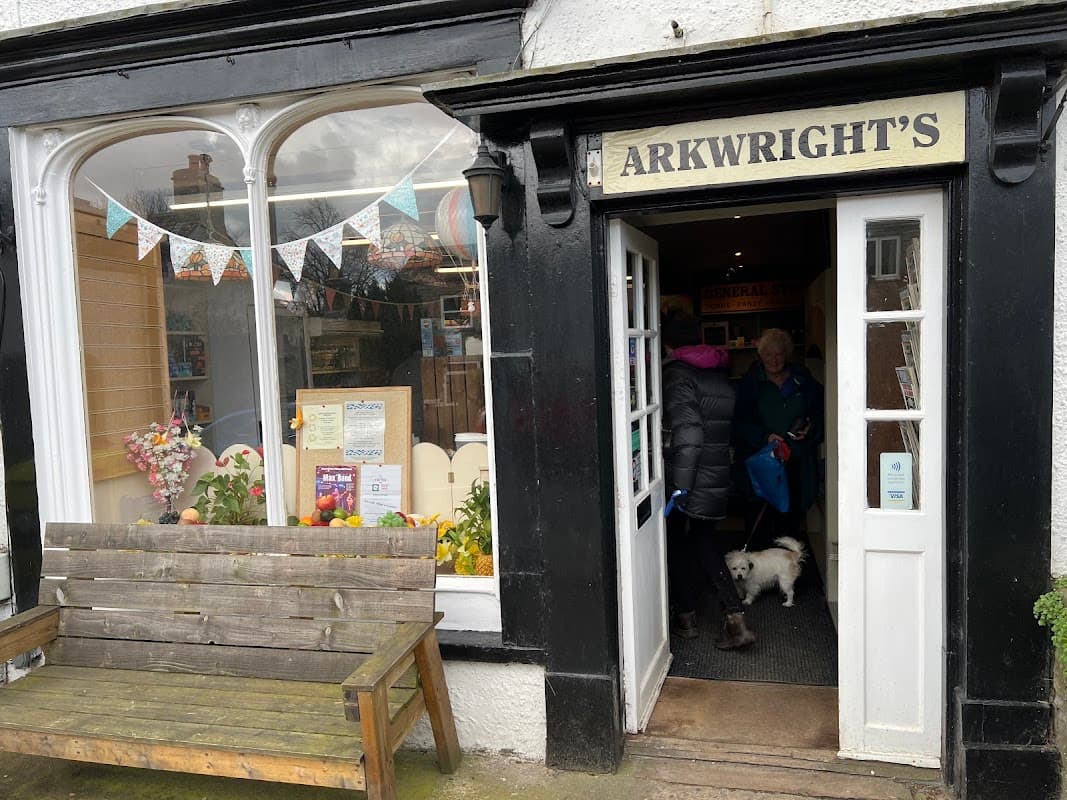 Quaint village shop entrance with a wooden bench, colorful bunting, and customers entering with a small dog.