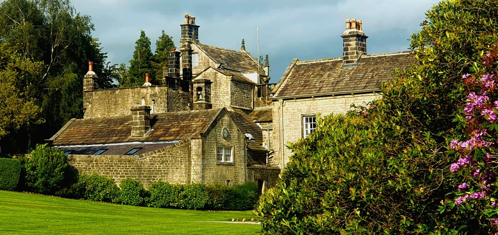 Historic stone building with multiple chimneys, surrounded by lush greenery and blooming shrubs under a cloudy sky.