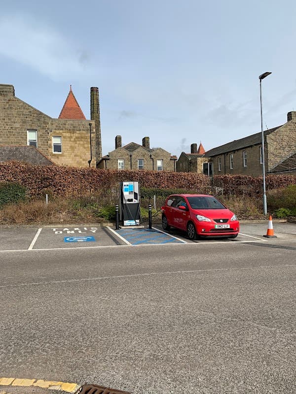 Parking area with a red car and an electric vehicle charging station, surrounded by stone buildings and greenery.