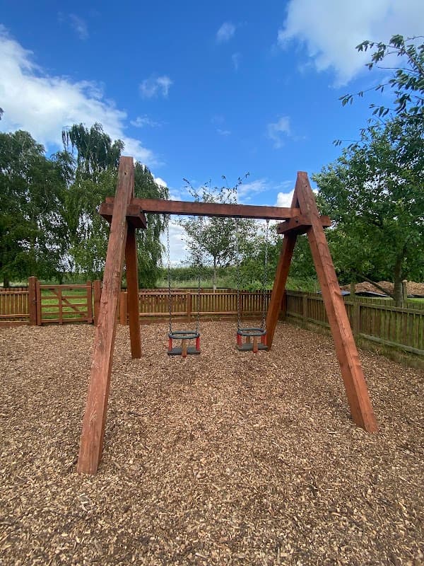 Wooden swing set with two swings, surrounded by a fenced area and lush greenery under a blue sky.