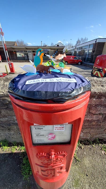 A red post box with a colorful wildlife-themed decoration on top, set in a parking area with buildings in the background.