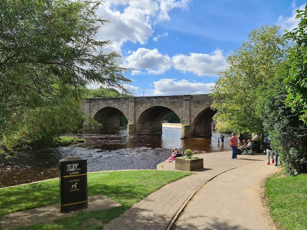 Wilderness Car Park by a river, with a stone bridge, trees, and people enjoying the scenic view on a sunny day.