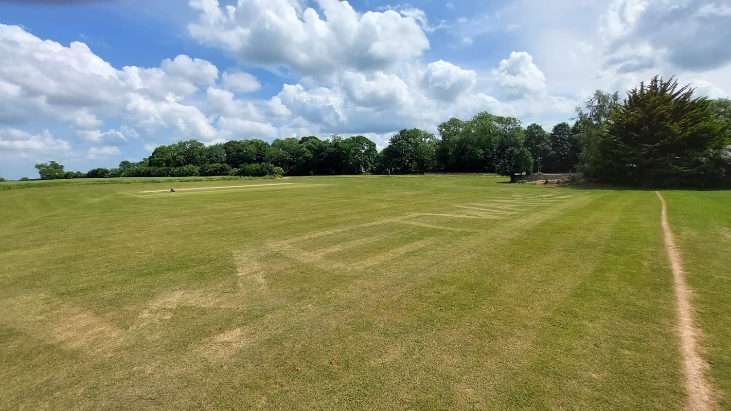 Cricket field with "WETWANG" mowed into the grass, surrounded by trees and a blue sky with fluffy clouds.