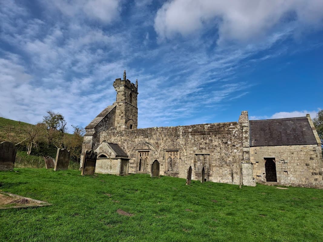 Medieval stone church with a tower, surrounded by grassy grounds and gravestones under a blue sky with clouds.