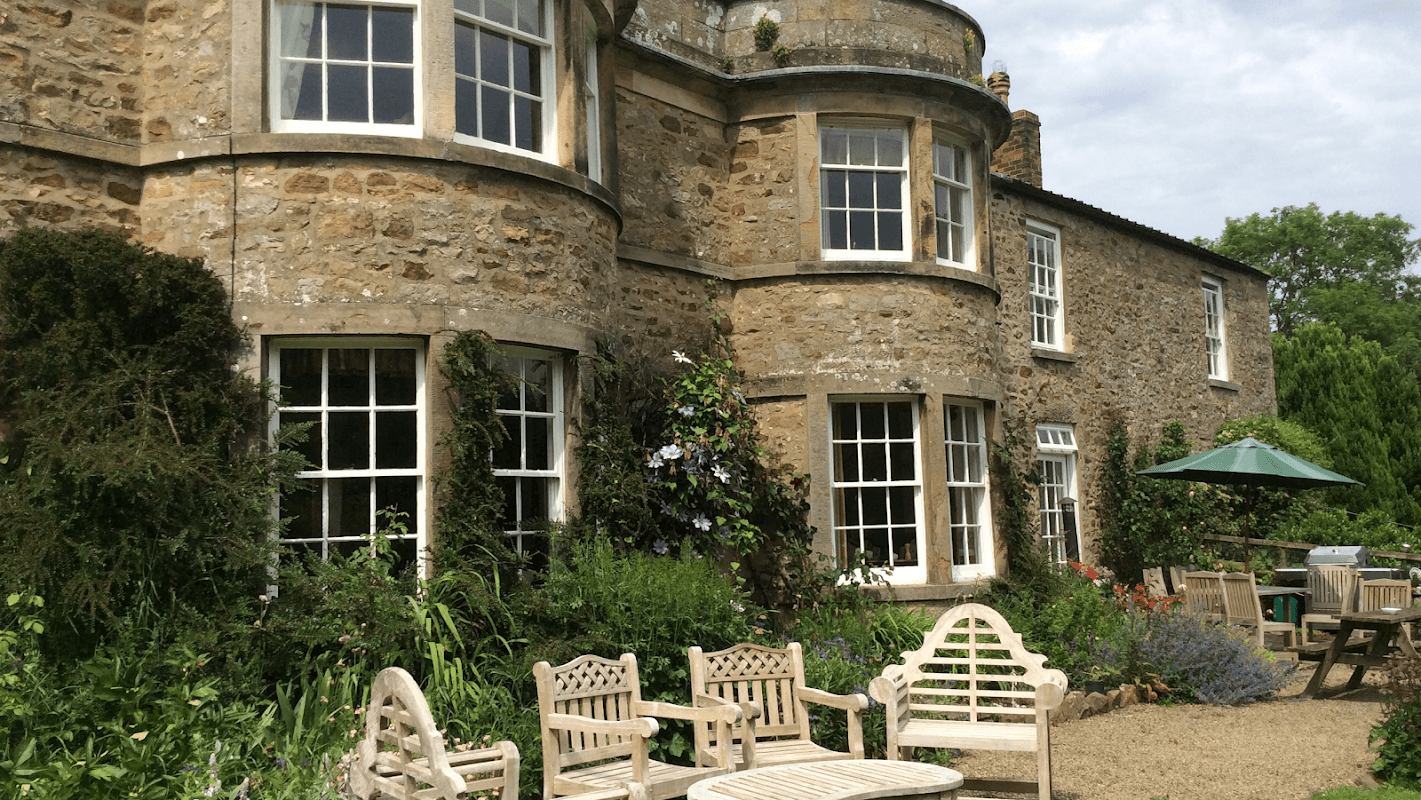 Stone farmhouse with large windows, surrounded by lush greenery and outdoor seating in a tranquil garden setting.