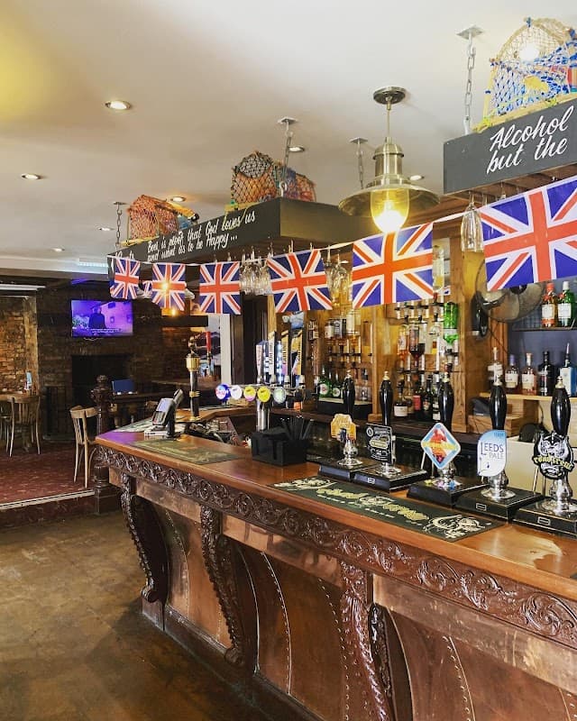 Wooden bar with British flags, shelves of spirits, and a TV screen in a cozy pub setting.