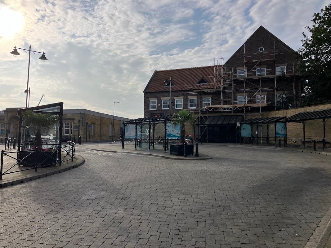 Whitby bus station with a cobblestone area, palm trees, and a building under scaffolding against a cloudy sky.