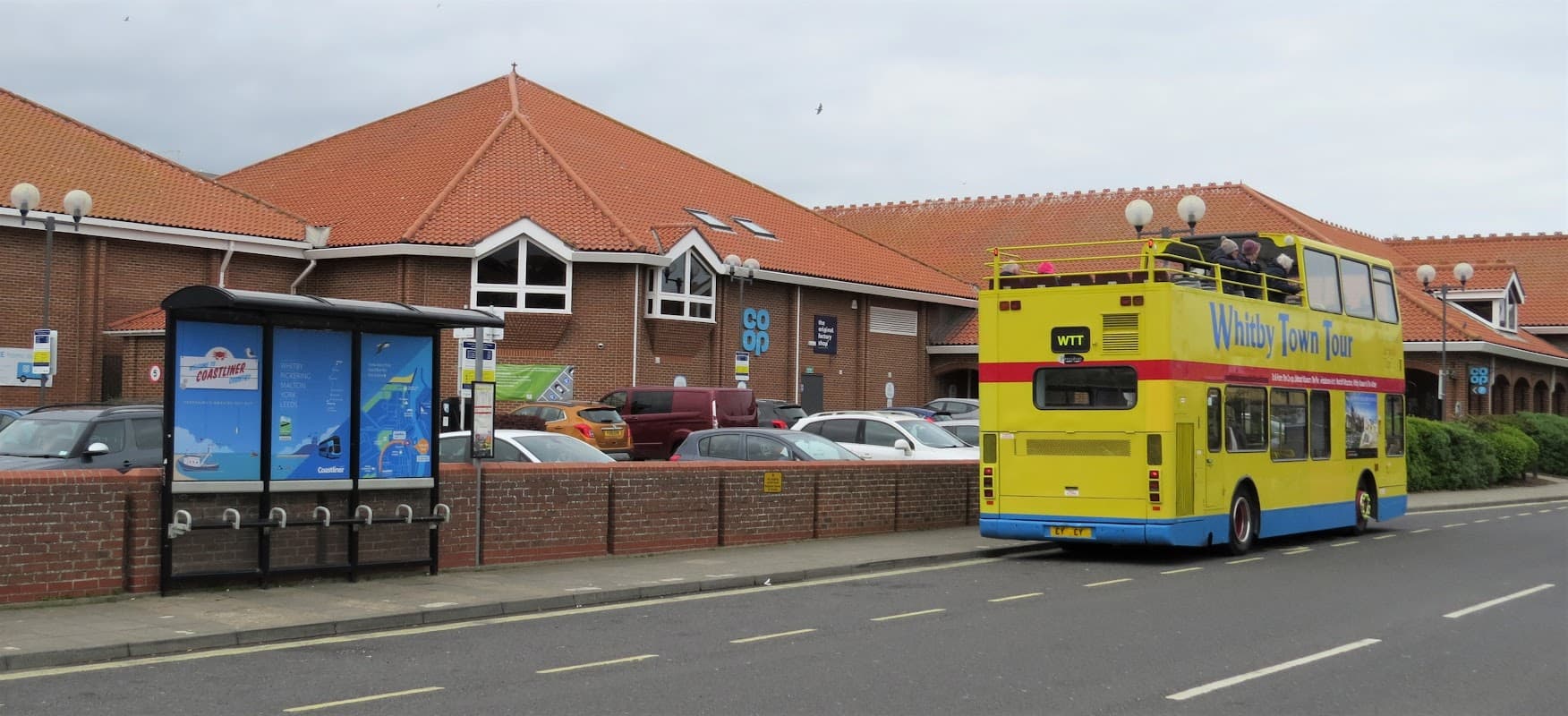 Bright yellow double-decker bus parked outside the Whitby Co-op, with a bus stop and cars in the background.