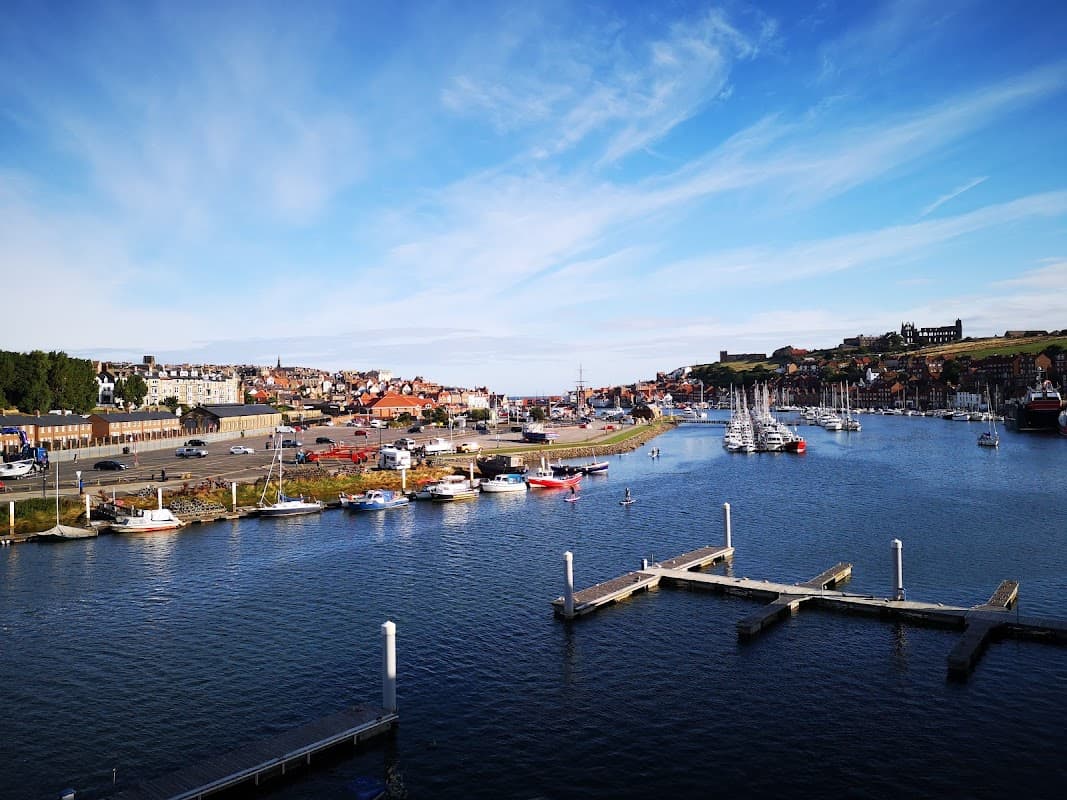 View of Whitby Coach Park with parked vehicles, boats in the harbor, and a scenic coastline under a blue sky.