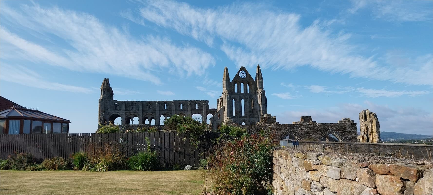 Whitby Holiday Cottages with lush greenery and the ruins of Whitby Abbey under a blue sky with scattered clouds.