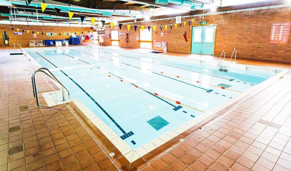 Indoor swimming pool with clear water, lane markings, and colorful bunting along the walls.
