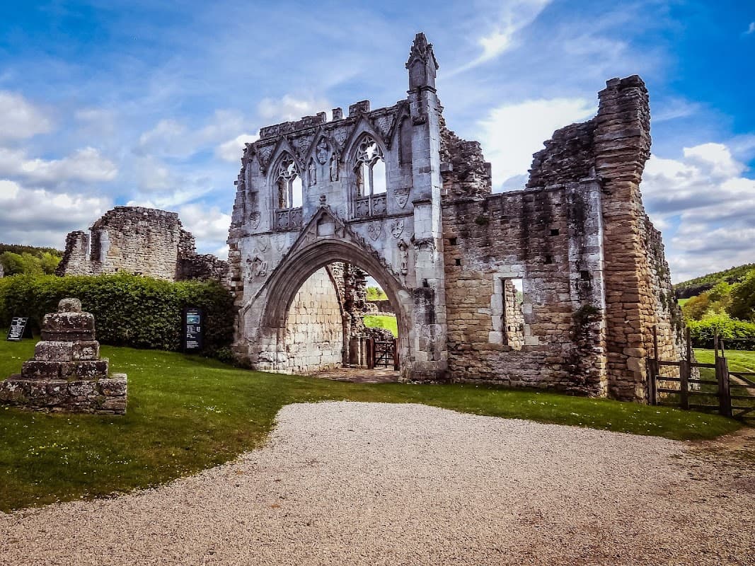 Ruins of Kirkham Priory with stone archway, grassy area, and trees under a blue sky with clouds.