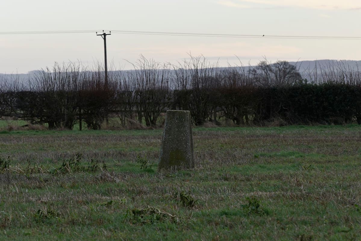 Trig point on a grassy field, surrounded by hedgerows and distant hills under a soft sky.
