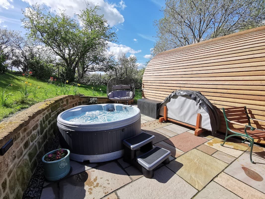 Hot tub with steps surrounded by greenery and trees, alongside a wooden pod structure in a sunny outdoor setting.