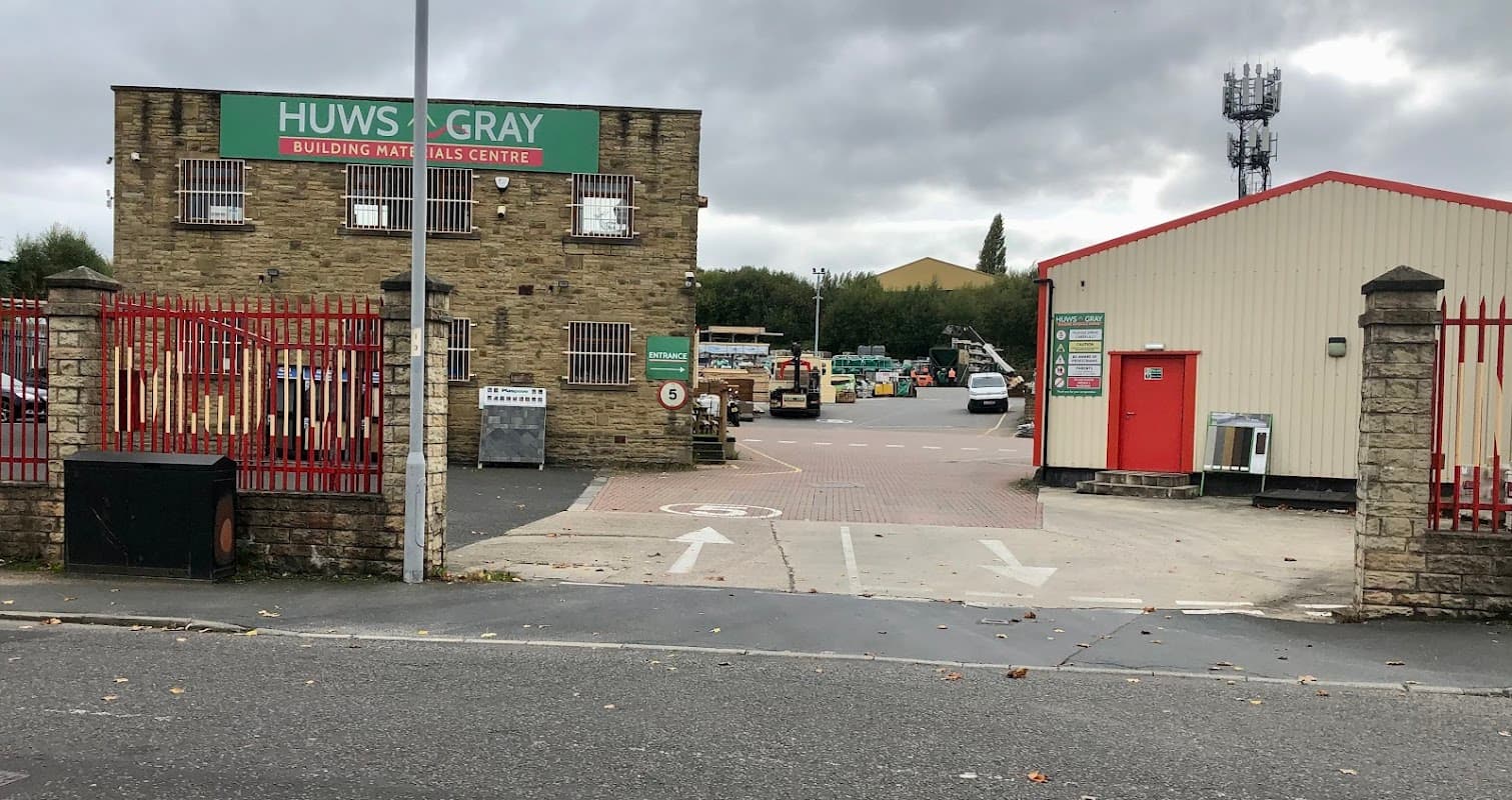 Huws Gray building materials center with red gates, stone building, and various vehicles in the parking lot.