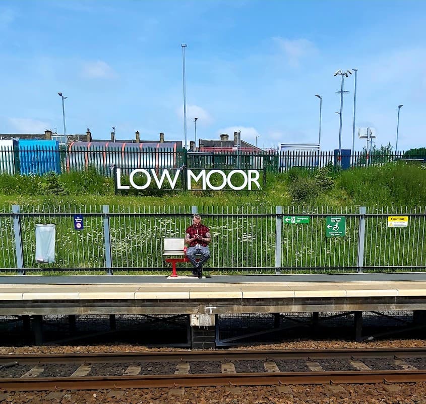 Low Moor station car park with a man sitting on a bench, greenery, and signage in the background.