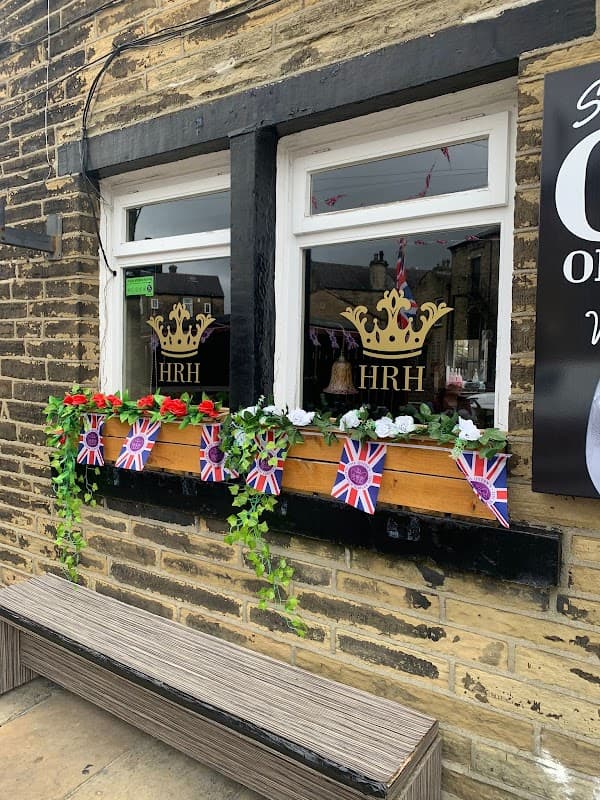 Bar windows adorned with flowers, Union Jack flags, and a crown logo, set against a stone wall in Wibsey, Yorkshire.