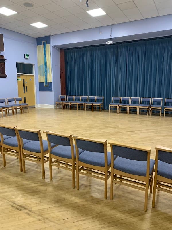 Interior of Wibsey Methodist Church featuring wooden chairs arranged in a circle on a wooden floor with blue curtains.
