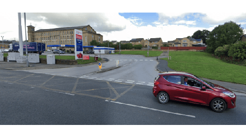 Red car driving on a road near a parking area and buildings in Wibsey, Yorkshire, with green grass and cloudy sky.
