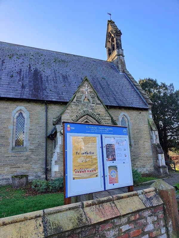 St Nicholas Church with a stone facade, a prominent steeple, and an information board in front against a clear blue sky.