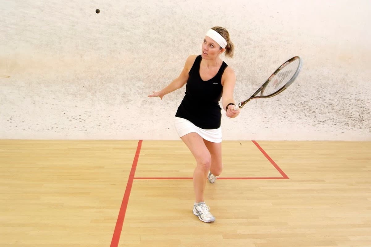 A woman in athletic wear plays squash, swinging a racquet in a court with a textured wall and wooden floor.