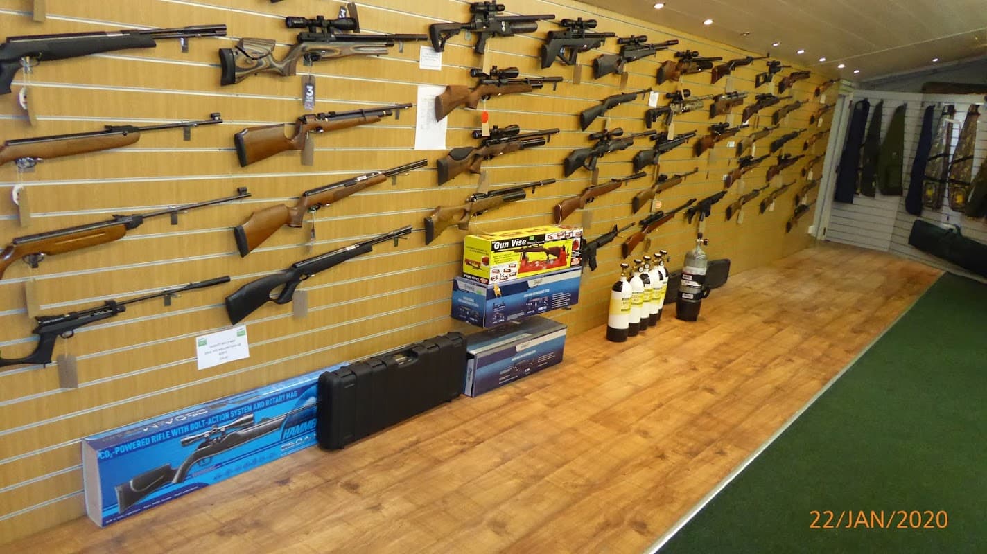 Wall display of various rifles and shotguns, with ammunition boxes and cases on the floor in a shop setting.
