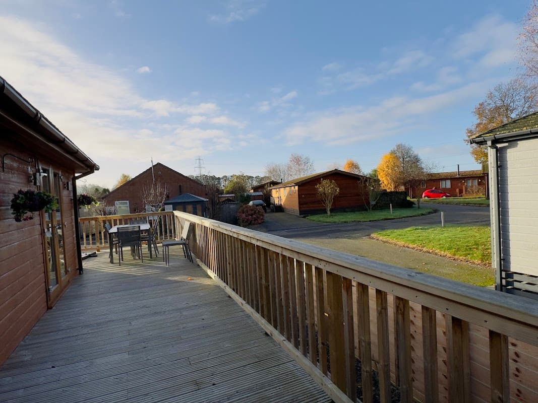 Wooden deck with outdoor seating, surrounded by lodges and trees under a clear blue sky in Wilberfoss, Yorkshire.