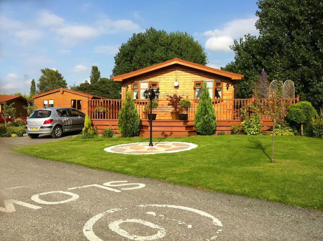 Wooden holiday cabin with a deck, surrounded by greenery and decorative plants, on a well-maintained grassy area.