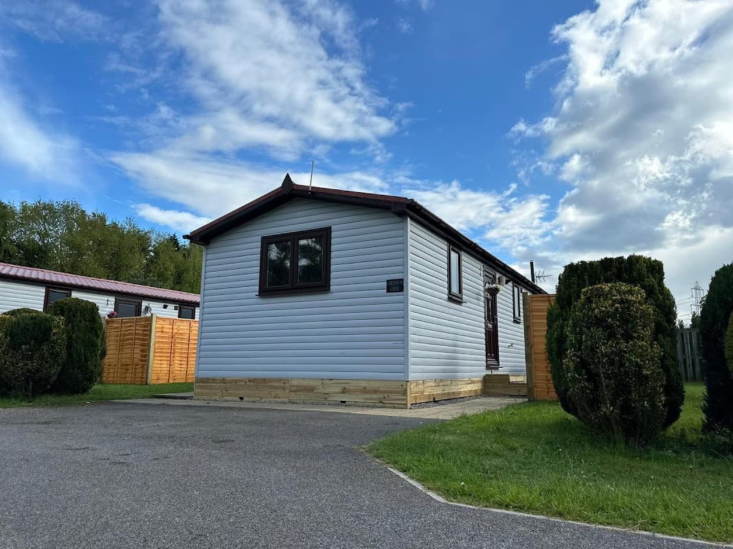 White holiday cabin with wooden steps, surrounded by greenery and wooden fencing under a partly cloudy sky.