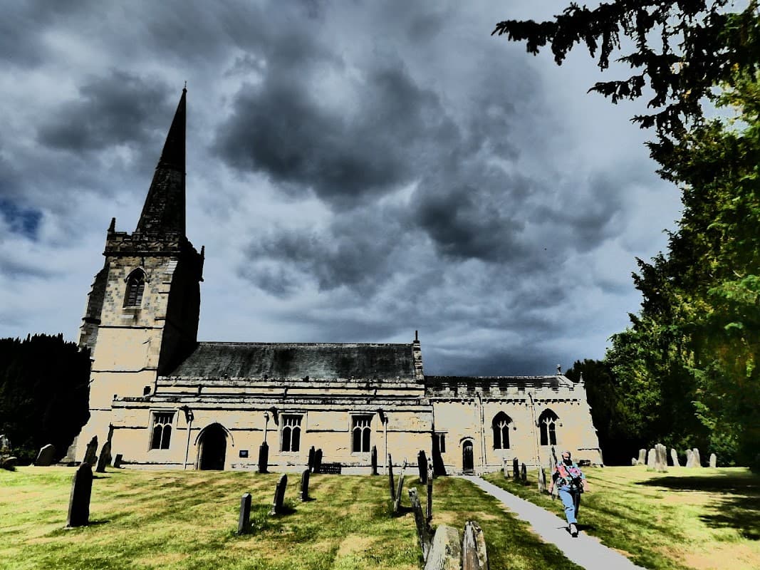 St Peter's Church with a tall spire, surrounded by gravestones and lush greenery under a dramatic cloudy sky.