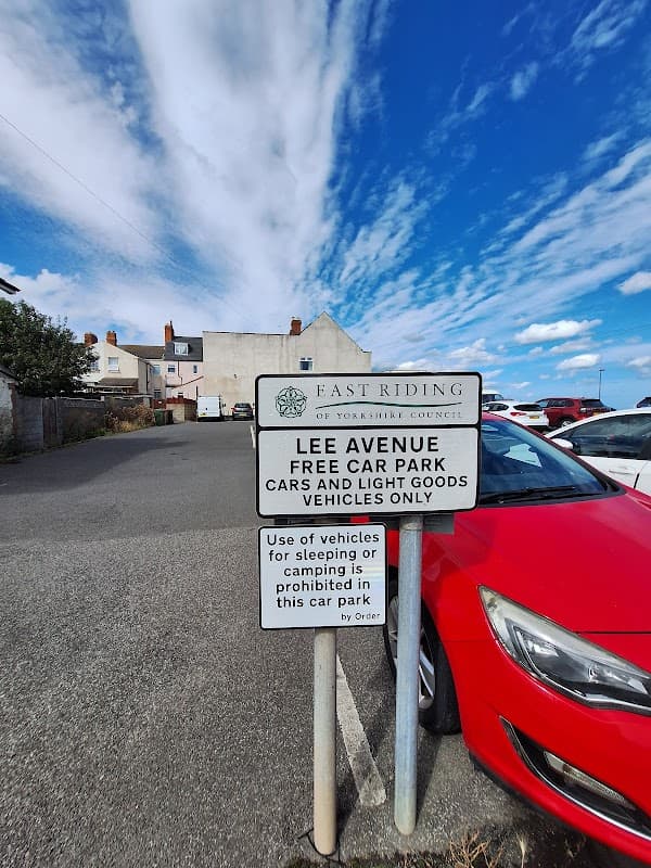 Sign for Lee Avenue Free Car Park, with a red car parked nearby and a cloudy blue sky above.