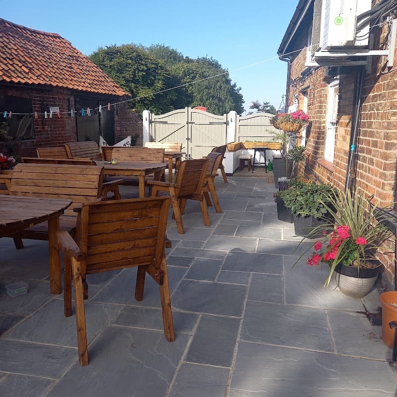 Outdoor seating area with wooden tables and chairs, stone flooring, and potted plants at The Anvil Arms in Wold Newton.