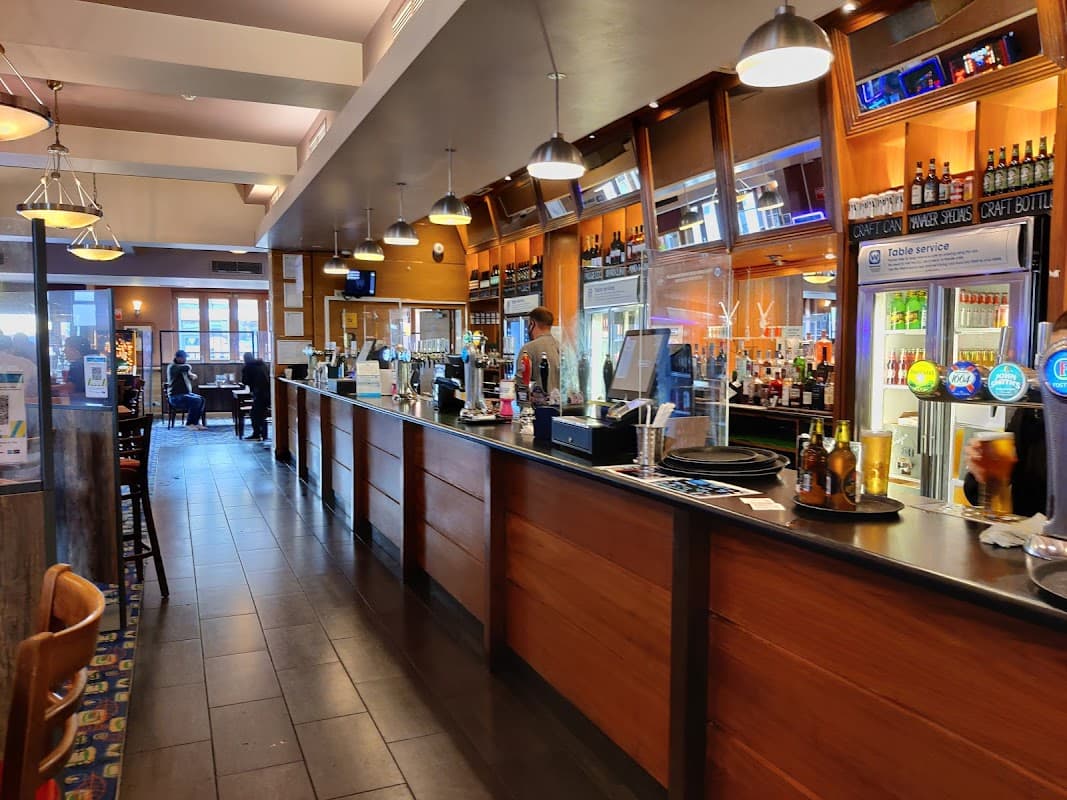 Modern bar area with wooden counters, drinks on display, and patrons seated in a cozy restaurant setting.