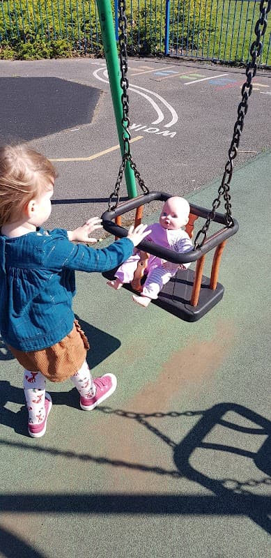 A child pushes a doll in a swing at a playground with colorful surfaces and a clear blue sky in Wombwell Skate Park.