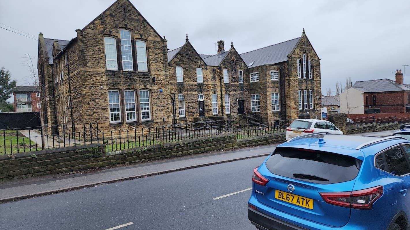 Historic stone building with large windows, surrounded by a low stone wall, parked cars on the street.