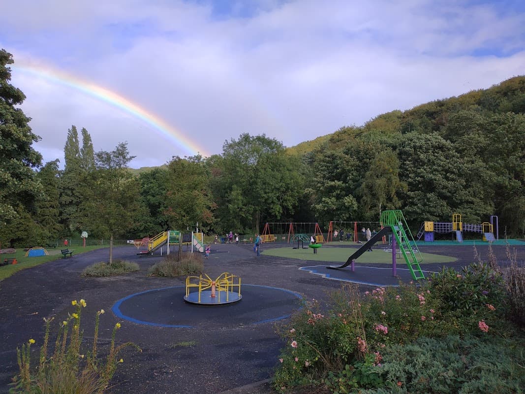 Colorful playground equipment under a rainbow, surrounded by green trees and blooming flowers in Graves Park, Woodseats.