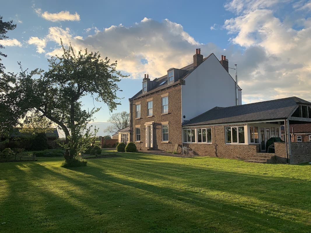 Luxury cottage with a spacious lawn, trees, and a two-story brick exterior under a blue sky with fluffy clouds.