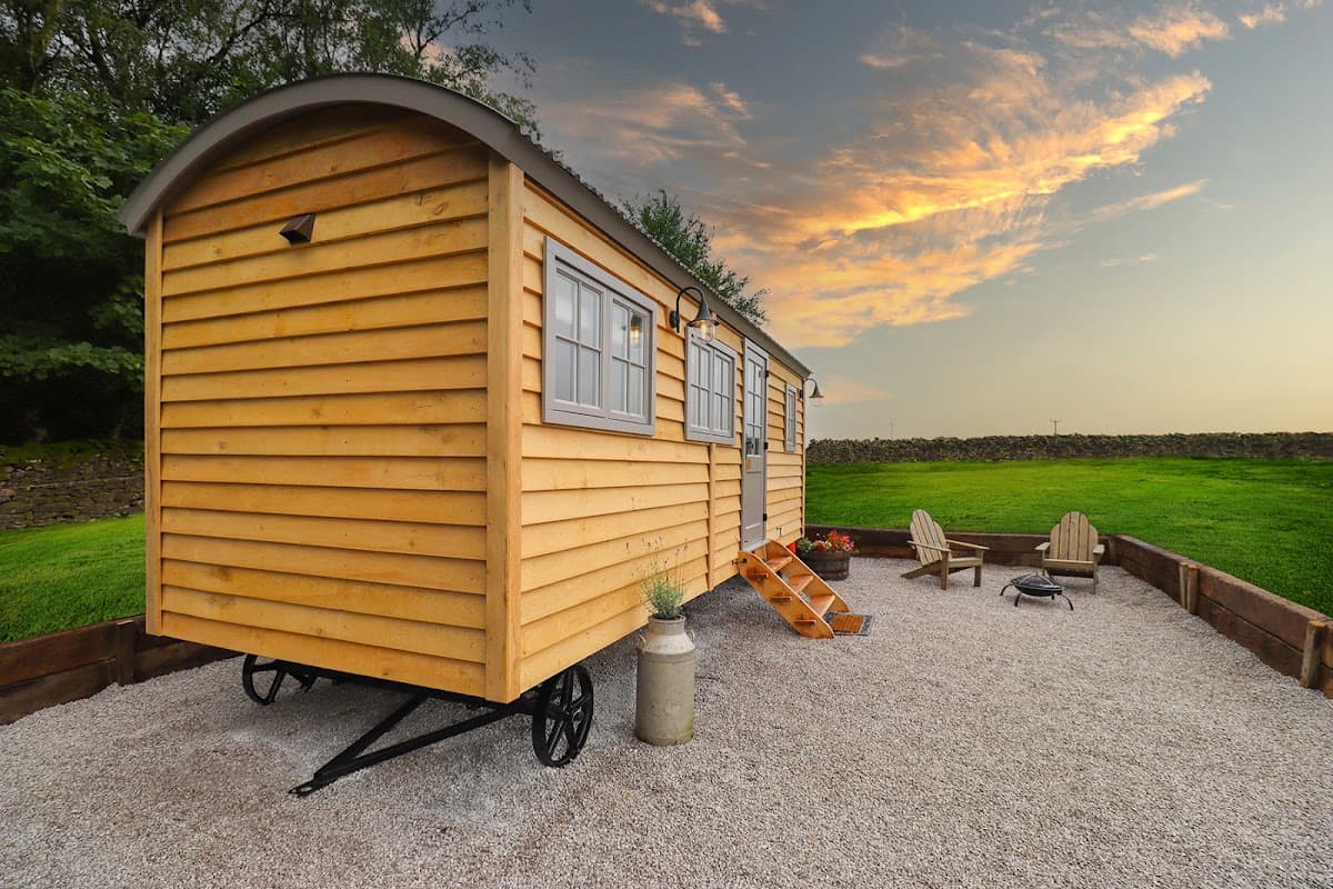 Yellow wooden hut on a gravel campsite, with a fire pit and Adirondack chairs, set against a sunset sky.