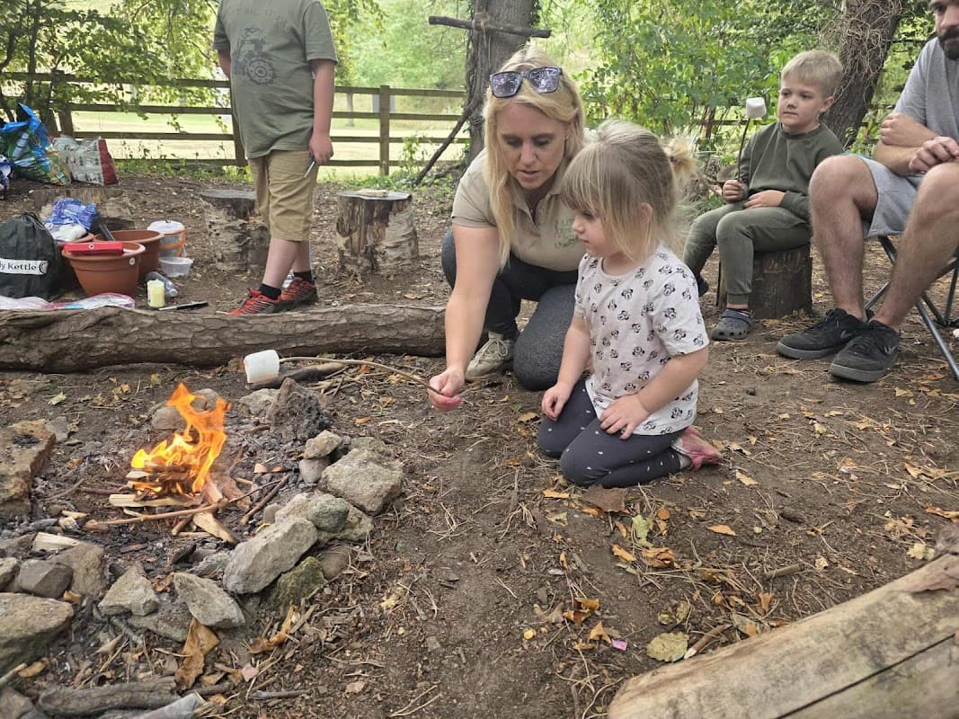 A woman teaches a young girl by a small campfire, surrounded by children and nature in a woodland setting.