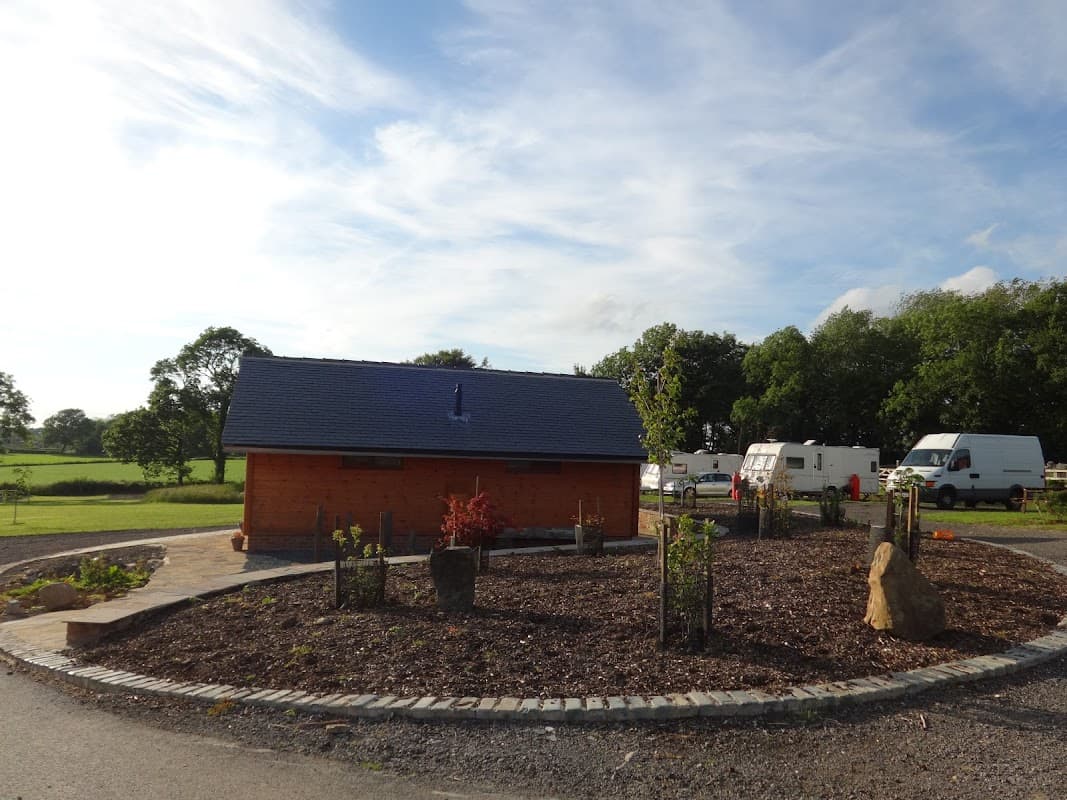 Wooden building with a circular garden, surrounded by green fields and parked caravans under a blue sky.
