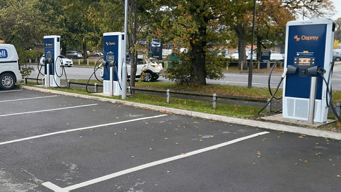 Four electric vehicle charging stations lined up in a parking lot, surrounded by trees and autumn leaves.
