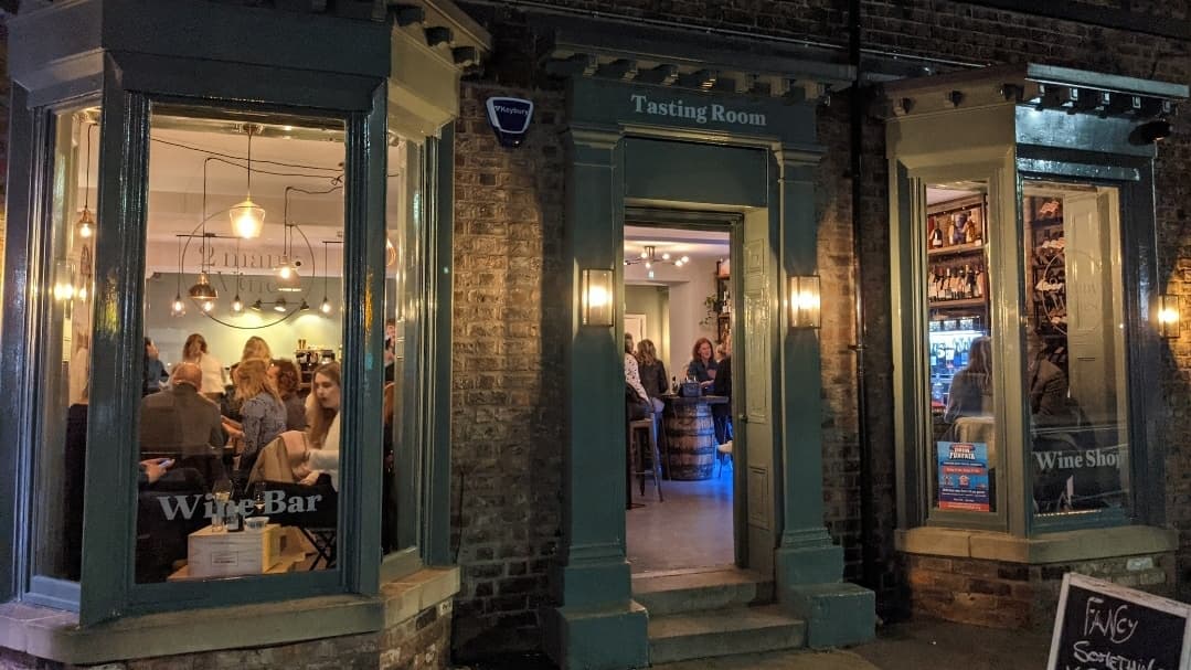 Entrance to "2 Many Wines" with large windows, showcasing a lively tasting room and wine displays.