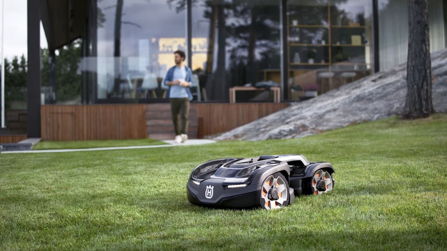 A black robotic lawn mower on green grass, with a modern building and a person in the background.