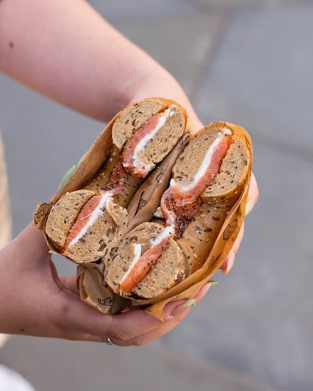 A person holding a halved bagel filled with cream cheese and smoked salmon, wrapped in paper.