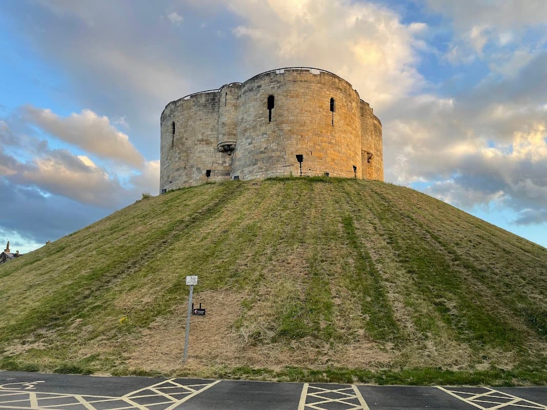 Castle on a grassy mound under a cloudy sky, with a car park and parking lines in the foreground.