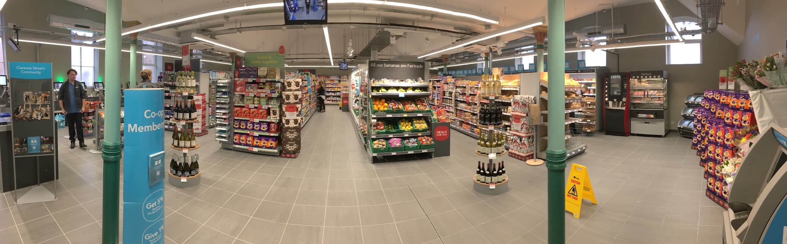 Brightly lit corner shop interior with aisles of groceries, drinks, and snacks, featuring signage and a clean layout.