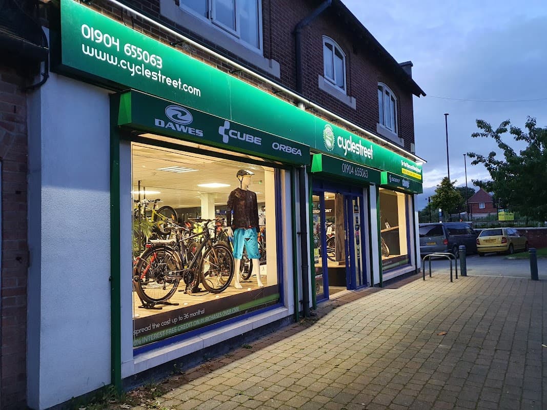 Bike shop exterior with illuminated display showcasing bicycles and cycling gear, green signage, and nearby parked cars.