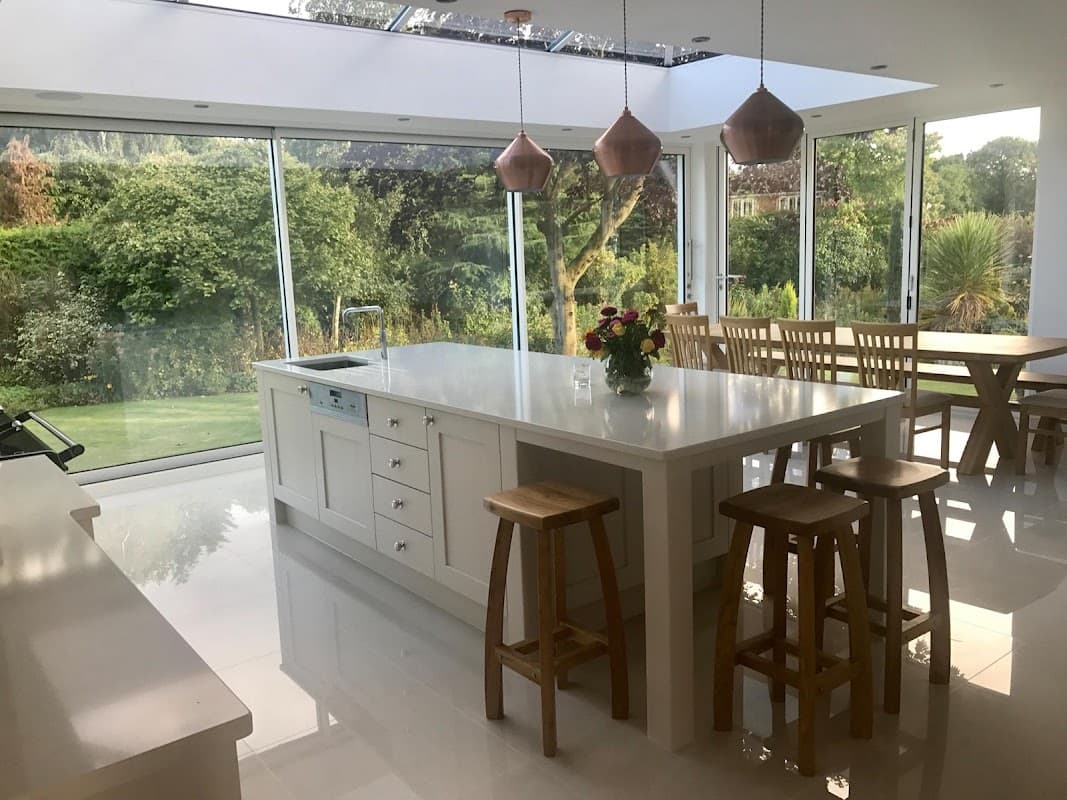 Bright, modern kitchen with white cabinetry, a large island, wooden stools, and glass walls overlooking a garden.