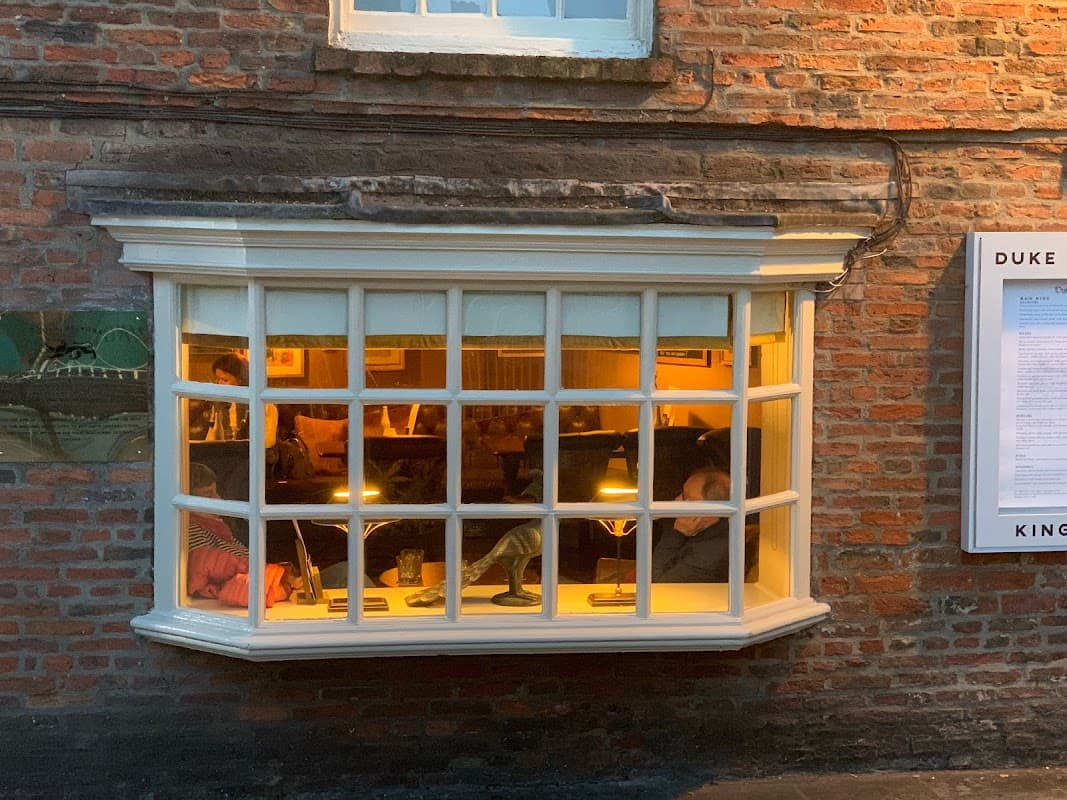 Cozy pub window with warm lighting, brick wall exterior, and patrons enjoying drinks inside.
